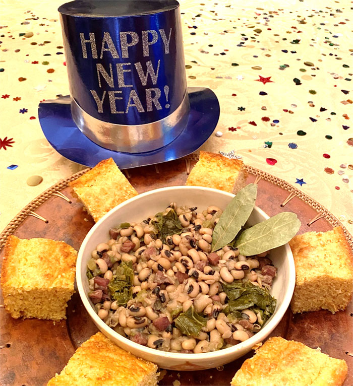 bowl of tan beans with black dots on top surrounded by corn bread squares and a paper top hat that says Happy New Year!
