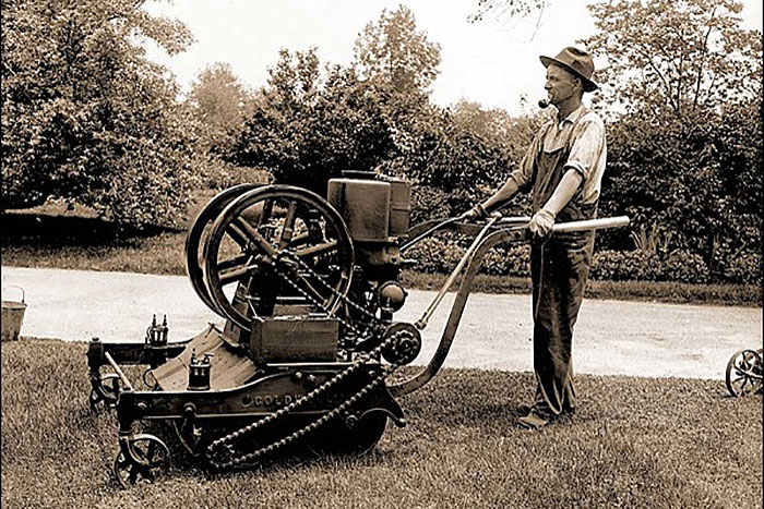 black and white image of man standing behind an early lawnmower